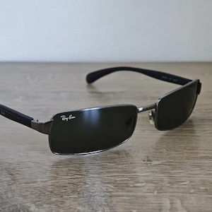 May include: Rectangular sunglasses with dark lenses and a silver frame. The Ray-Ban logo is visible on the lens. The temples are black and curve slightly. The sunglasses are resting on a wooden surface.