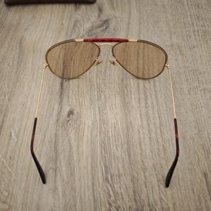 K&ouml;nnte beinhalten: Sonnenbrille im Aviator-Stil mit goldfarbenem Rahmen und braunen Gl&auml;sern. Der obere Steg ist dunkelrot und die B&uuml;gel haben ein Schildpattmuster. Die Sonnenbrille liegt auf einem Holzgrund.