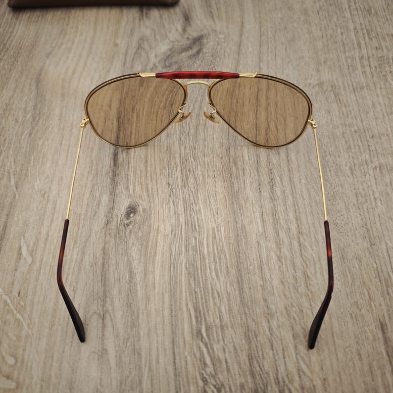 K&ouml;nnte beinhalten: Sonnenbrille im Aviator-Stil mit goldfarbenem Rahmen und braunen Gl&auml;sern. Der obere Steg ist dunkelrot und die B&uuml;gel haben ein Schildpattmuster. Die Sonnenbrille liegt auf einem Holzgrund.