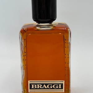 Könnte beinhalten: Eine Vintage-Cologne-Flasche mit schwarzem Verschluss und einem rechteckigen, bernsteinfarbenen Glaskörper. Das Etikett trägt die Aufschrift "BRAGGI" in weißen Buchstaben auf schwarzem Grund. Die Flasche hat ein strukturiertes Design an den Seiten.