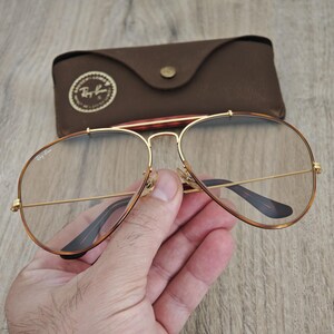 K&ouml;nnte beinhalten: Goldgerahmte Aviator-Brille mit klaren Gl&auml;sern. Die Brille hat einen Doppelsteg und braune B&uuml;gel. Im Hintergrund befindet sich ein braunes Lederetui mit Knopfverschluss und Logo.