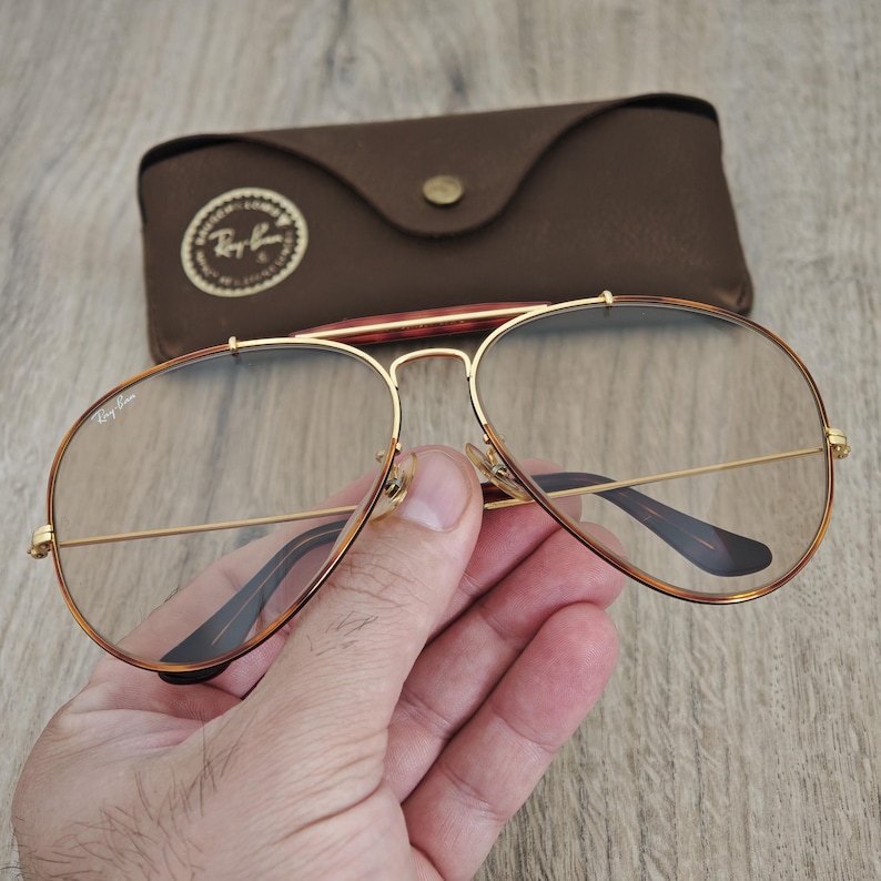 K&ouml;nnte beinhalten: Goldgerahmte Aviator-Brille mit klaren Gl&auml;sern. Die Brille hat einen Doppelsteg und braune B&uuml;gel. Im Hintergrund befindet sich ein braunes Lederetui mit Knopfverschluss und Logo.