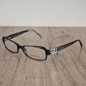 May include: Black rectangular spectacles featuring a textured frame finish and the Versace logo printed on the right lens. The temples display a silver-toned metal accent with a Greek key pattern embedded with small clear crystals and the Versace name.
