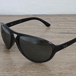 Puede incluir: Gafas de sol negras con un dise&ntilde;o curvo. Las gafas tienen lentes oscuras y una montura negra brillante. El logotipo de Ray-Ban es visible en la patilla derecha. Est&aacute;n sobre una superficie de madera.