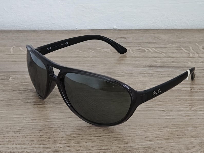 Puede incluir: Gafas de sol negras con un dise&ntilde;o curvo. Las gafas tienen lentes oscuras y una montura negra brillante. El logotipo de Ray-Ban es visible en la patilla derecha. Est&aacute;n sobre una superficie de madera.