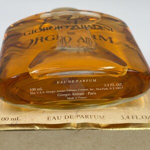 May include: A clear glass perfume bottle filled with amber liquid, featuring the text Gio de Giorgio Armani. The base displays Eau de Parfum, 100 mL, 3.4 fl. oz., and Made in France. The bottle rests on a gold-colored cardboard box.
