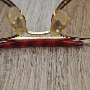 K&ouml;nnte beinhalten: Nahaufnahme einer Ray-Ban-Brille. Die Brille hat einen goldfarbenen Rahmen mit einem dunkelrotbraunen B&uuml;gel. Die Nasenpads sind hellbeige. Der Text "B&L RAY-BAN 62 14" ist auf dem Rahmen zu sehen.