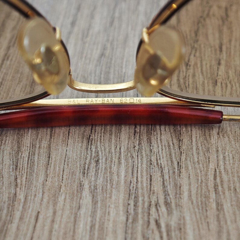 K&ouml;nnte beinhalten: Nahaufnahme einer Ray-Ban-Brille. Die Brille hat einen goldfarbenen Rahmen mit einem dunkelrotbraunen B&uuml;gel. Die Nasenpads sind hellbeige. Der Text "B&L RAY-BAN 62 14" ist auf dem Rahmen zu sehen.