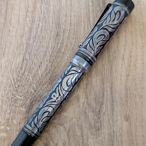 以下が含まれることがあります： 黒とシルバーの万年筆。装飾的な彫刻が施された花柄模様。ペンにはシルバーのクリップとアクセント、黒いペン先が付いています。木製の表面にペンが置かれています。