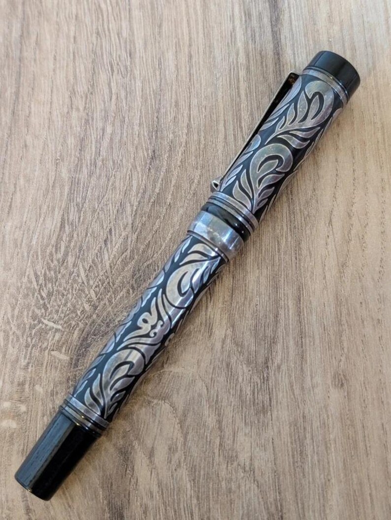 以下が含まれることがあります： 黒とシルバーの万年筆。装飾的な彫刻が施された花柄模様。ペンにはシルバーのクリップとアクセント、黒いペン先が付いています。木製の表面にペンが置かれています。