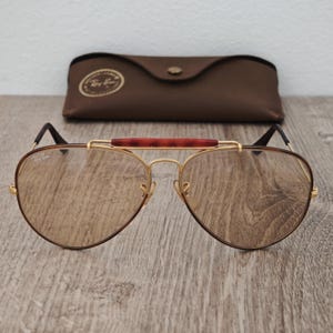 K&ouml;nnte beinhalten: Braune Aviator-Sonnenbrille mit goldenem Rahmen und braunem Lederetui. Die Sonnenbrille hat einen braunen Steg und braune Gl&auml;ser. Das Etui ist dunkelbraun und hat einen Knopfverschluss.