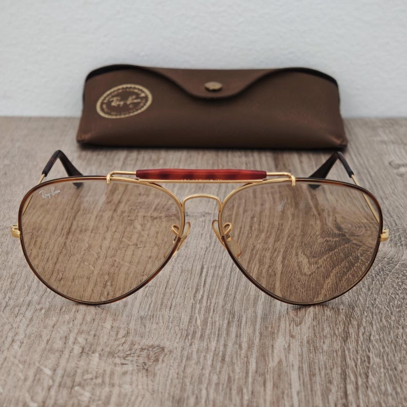 K&ouml;nnte beinhalten: Braune Aviator-Sonnenbrille mit goldenem Rahmen und braunem Lederetui. Die Sonnenbrille hat einen braunen Steg und braune Gl&auml;ser. Das Etui ist dunkelbraun und hat einen Knopfverschluss.