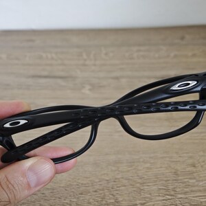 May include: Black eyeglasses with a distinctive design. The frames feature the Oakley logo in white on the temples. The glasses have a sleek, modern aesthetic.