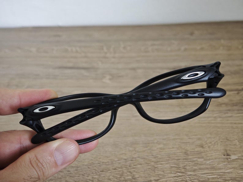 May include: Black eyeglasses with a distinctive design. The frames feature the Oakley logo in white on the temples. The glasses have a sleek, modern aesthetic.