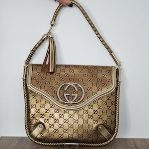 May include: A gold-toned shoulder bag with a repeating logo pattern. The bag features a silver-colored logo emblem, a braided strap, and a tassel. The bag's design includes gold trim and a flap closure.