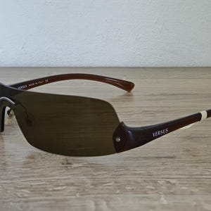 May include: Brown Versus sunglasses with a shield-style lens. The sunglasses have dark brown frames and arms with the Versus logo. The lenses are a gradient brown color. The sunglasses are made in Italy.
