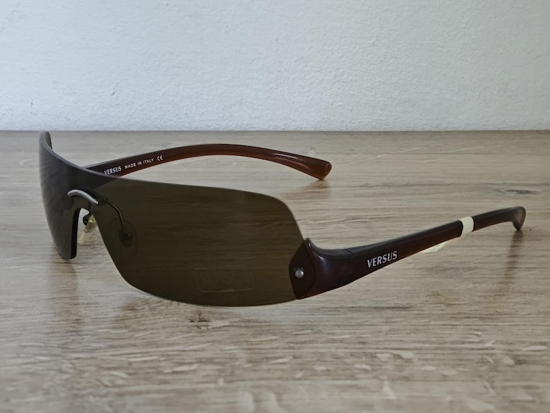 May include: Brown Versus sunglasses with a shield-style lens. The sunglasses have dark brown frames and arms with the Versus logo. The lenses are a gradient brown color. The sunglasses are made in Italy.