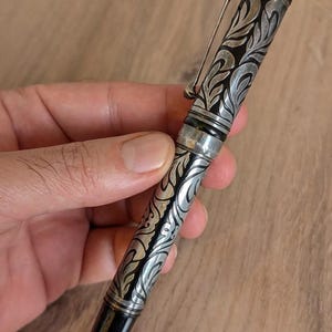 以下が含まれることがあります： 銀と黒の万年筆。装飾的なフローラルデザイン。ペンはシルバーのクリップと黒のペン先を備えています。ペンの本体には、詳細な渦巻き模様が刻まれています。ペンは手で持たれています。