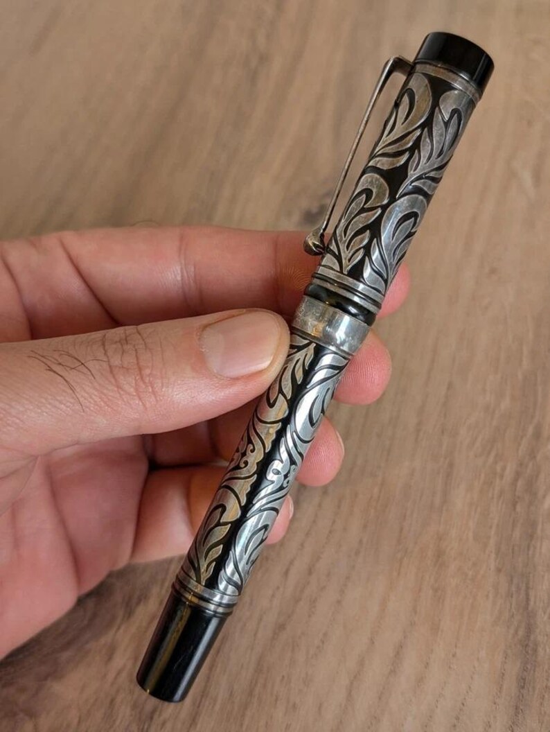 以下が含まれることがあります： 銀と黒の万年筆。装飾的なフローラルデザイン。ペンはシルバーのクリップと黒のペン先を備えています。ペンの本体には、詳細な渦巻き模様が刻まれています。ペンは手で持たれています。