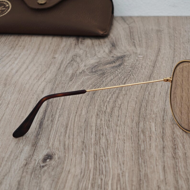 K&ouml;nnte beinhalten: Goldfarbene Pilotenbrille mit braunen Gl&auml;sern. Die B&uuml;gel sind goldfarben mit dunkelbraunen Ohrenb&uuml;geln. Im Hintergrund ist ein braunes Lederetui zu sehen. Die Sonnenbrille liegt auf einer Holzoberfl&auml;che.