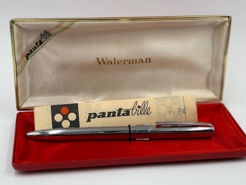 K&ouml;nnte beinhalten: Ein silberner Waterman-Stift in einer roten und goldenen Schatulle. Die offene Schatulle zeigt den Stift, einen Papiereinleger mit dem Text "panta bille" und das Wort "Waterman" auf dem Deckel.