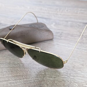 K&ouml;nnte beinhalten: Sonnenbrille mit goldfarbenem Rahmen und dunkelgr&uuml;nen Gl&auml;sern sowie passendem Etui. Die Sonnenbrille hat ein klassisches Aviator-Design mit einem d&uuml;nnen Goldrahmen und einem gebogenen oberen B&uuml;gel. Das Etui ist hellbraun.