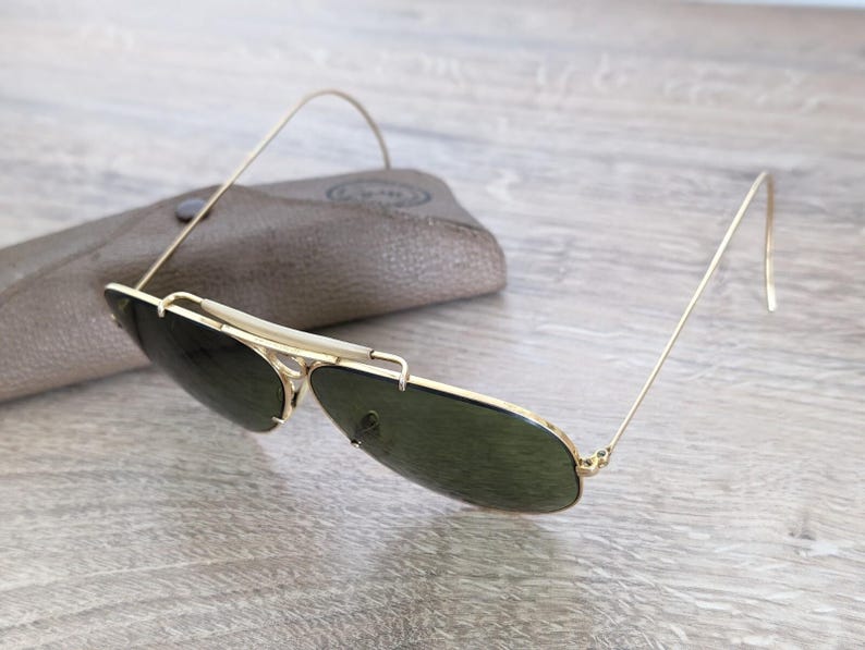 K&ouml;nnte beinhalten: Sonnenbrille mit goldfarbenem Rahmen und dunkelgr&uuml;nen Gl&auml;sern sowie passendem Etui. Die Sonnenbrille hat ein klassisches Aviator-Design mit einem d&uuml;nnen Goldrahmen und einem gebogenen oberen B&uuml;gel. Das Etui ist hellbraun.