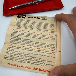K&ouml;nnte beinhalten: Silberner Stift mit schwarzem Band und blauen Akzenten, pr&auml;sentiert in einem roten Etui. Dem Stift liegt ein Papier mit Anweisungen und dem Text "Pantabille, une cr&eacute;ation Jif Waterman" bei.