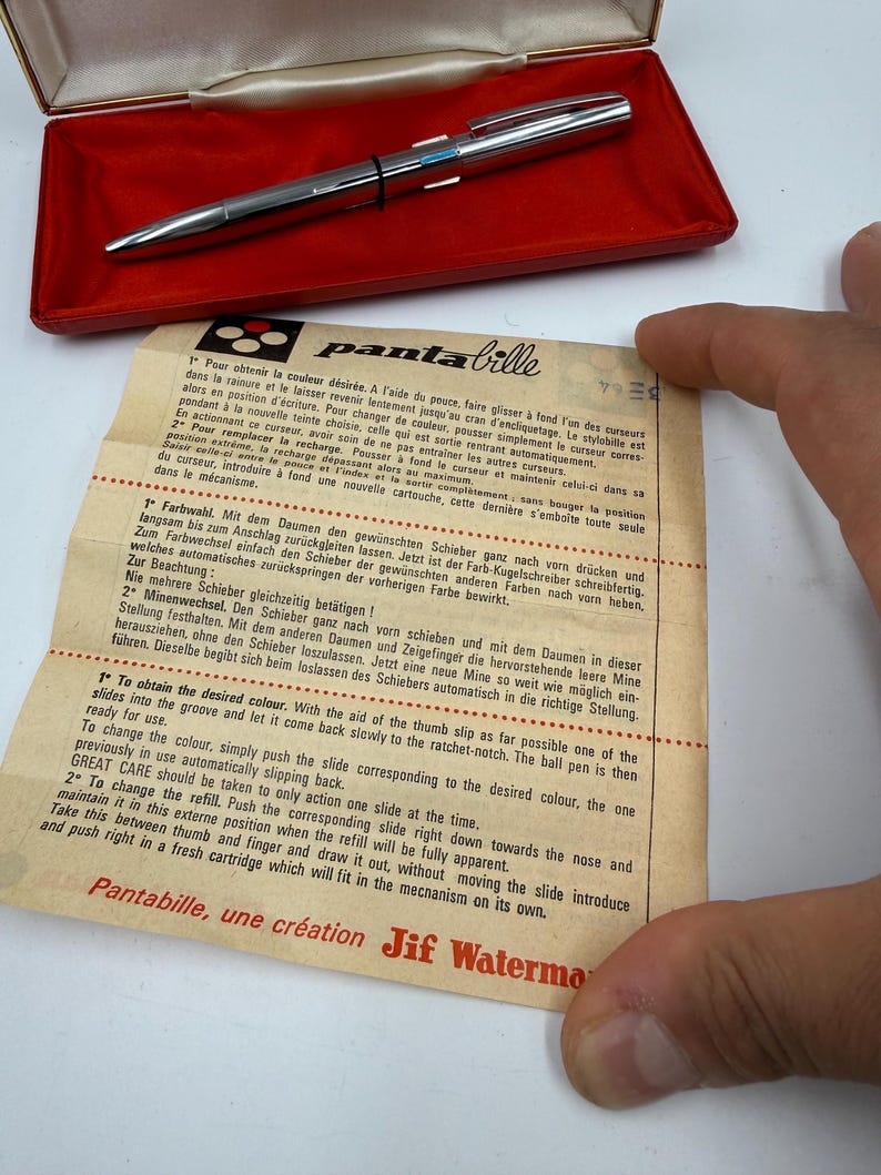 K&ouml;nnte beinhalten: Silberner Stift mit schwarzem Band und blauen Akzenten, pr&auml;sentiert in einem roten Etui. Dem Stift liegt ein Papier mit Anweisungen und dem Text "Pantabille, une cr&eacute;ation Jif Waterman" bei.