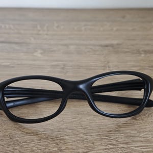 May include: Black eyeglasses with a rounded frame design. The glasses are made of a matte material and are resting on a wooden surface. The frames have a thick, solid appearance.
