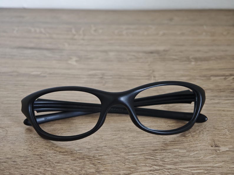 May include: Black eyeglasses with a rounded frame design. The glasses are made of a matte material and are resting on a wooden surface. The frames have a thick, solid appearance.