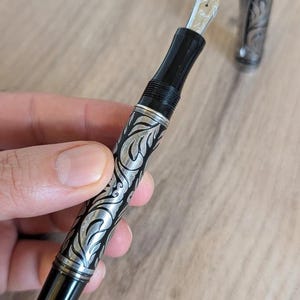 以下が含まれることがあります： 黒とシルバーの万年筆。 複雑な葉のような彫刻が施されています。 ペンはシルバーのペン先と、同様のディテールが施されたお揃いのキャップが特徴です。 明るい木製の表面に置かれています。