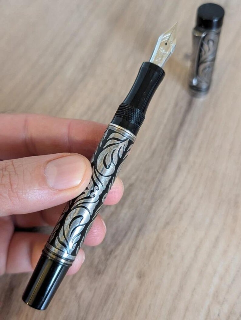 以下が含まれることがあります： 黒とシルバーの万年筆。 複雑な葉のような彫刻が施されています。 ペンはシルバーのペン先と、同様のディテールが施されたお揃いのキャップが特徴です。 明るい木製の表面に置かれています。