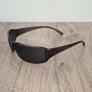 Puede incluir: Gafas de sol de color marr&oacute;n oscuro transl&uacute;cido con un dise&ntilde;o de montura envolvente y lentes tintadas oscuras. Las patillas de las gafas muestran la palabra VERSUS grabada en una fuente de letras may&uacute;sculas negritas en los laterales.