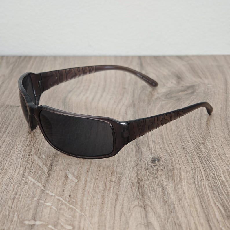 Puede incluir: Gafas de sol de color marr&oacute;n oscuro transl&uacute;cido con un dise&ntilde;o de montura envolvente y lentes tintadas oscuras. Las patillas de las gafas muestran la palabra VERSUS grabada en una fuente de letras may&uacute;sculas negritas en los laterales.