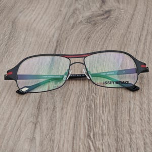 Può includere: Occhiali Issey Miyake neri con una striscia rossa sulla parte superiore della montatura. Gli occhiali hanno una forma rettangolare con una sottile montatura in metallo. Le lenti sono trasparenti e il nome del marchio è stampato sulla lente destra.