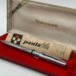 K&ouml;nnte beinhalten: Ein silberner Waterman panta bille Stift in einer rot samtgef&uuml;tterten Box. Der Stift hat ein schwarzes Band und einen blauen Akzent. Die Box ist cremefarben mit dem Wort "Waterman" in goldener Schrift. Der Deckel tr&auml;gt die Aufschrift "panta bille".