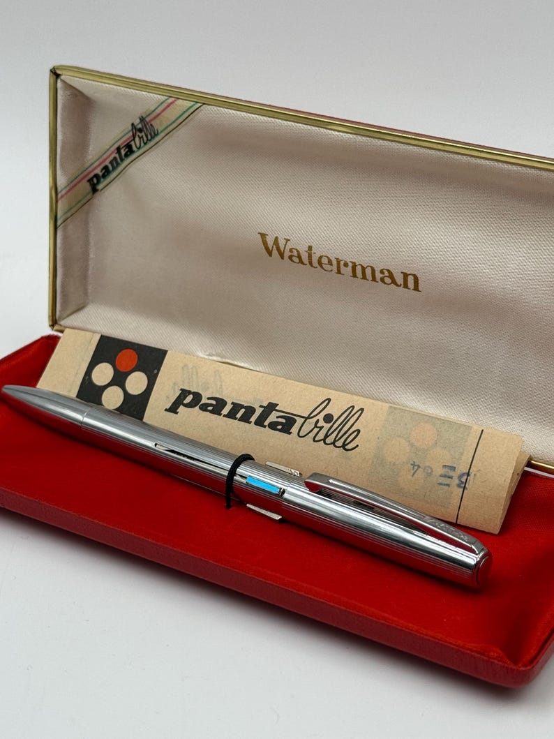 K&ouml;nnte beinhalten: Ein silberner Waterman panta bille Stift in einer rot samtgef&uuml;tterten Box. Der Stift hat ein schwarzes Band und einen blauen Akzent. Die Box ist cremefarben mit dem Wort "Waterman" in goldener Schrift. Der Deckel tr&auml;gt die Aufschrift "panta bille".