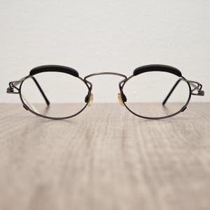 以下が含まれることがあります： ダークメタルフレームとクリアレンズの楕円形の眼鏡。 眼鏡には黒いパッド入りのレストと小さなノーズパッドが付いています。 デザインはミニマリストでモダンで、機能性とスタイルに重点を置いています。