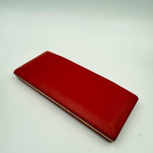 K&ouml;nnte beinhalten: Ein rechteckiges, rotes Etui mit strukturierter Oberfl&auml;che und d&uuml;nnem, silberfarbenem Rand. Das Etui ist geschlossen und scheint aus einem lederartigen Material zu bestehen. Der Hintergrund ist schlicht wei&szlig;.