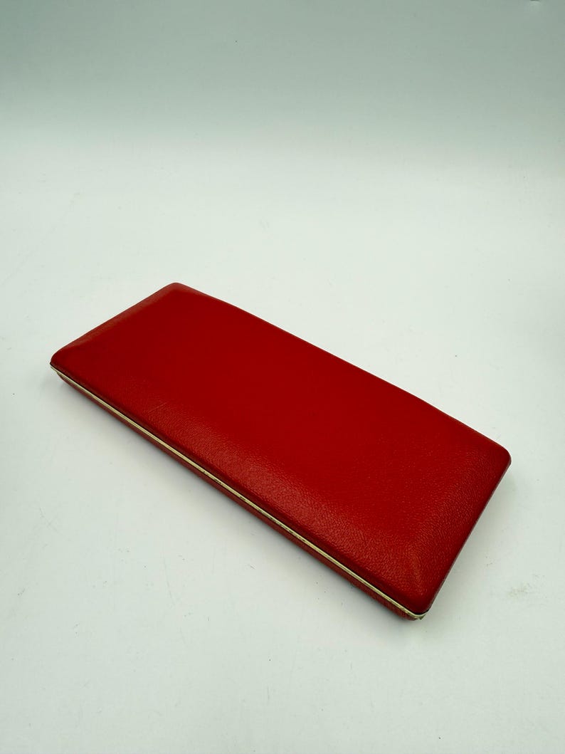 K&ouml;nnte beinhalten: Ein rechteckiges, rotes Etui mit strukturierter Oberfl&auml;che und d&uuml;nnem, silberfarbenem Rand. Das Etui ist geschlossen und scheint aus einem lederartigen Material zu bestehen. Der Hintergrund ist schlicht wei&szlig;.