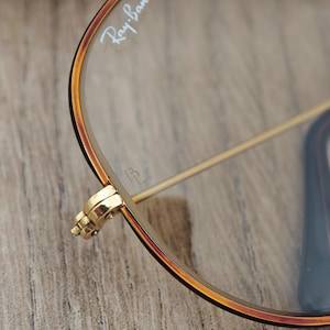 K&ouml;nnte beinhalten: Nahaufnahme einer Ray-Ban-Brille. Die Brille hat einen goldenen Rahmen mit einem braunen und schwarzen Rand. Das Ray-Ban-Logo ist auf dem Glas sichtbar. Das Scharnier ist goldfarben und detailliert. Die Brille liegt auf einer Holzoberfl&auml;che.