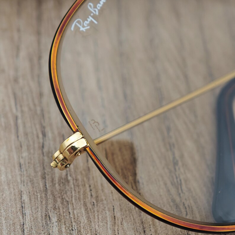 K&ouml;nnte beinhalten: Nahaufnahme einer Ray-Ban-Brille. Die Brille hat einen goldenen Rahmen mit einem braunen und schwarzen Rand. Das Ray-Ban-Logo ist auf dem Glas sichtbar. Das Scharnier ist goldfarben und detailliert. Die Brille liegt auf einer Holzoberfl&auml;che.