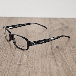 Peut inclure: Lunettes de vue gris foncé à motif écaille de tortue avec des montures rectangulaires. Les branches présentent un logo Versace argenté et un motif décoratif de petits cristaux incrustés. Le nom Versace est imprimé sur le verre droit.