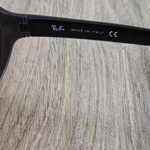 Puede incluir: Primer plano de la patilla de unas gafas de sol negras. El texto "Ray-Ban" est&aacute; impreso en blanco, junto con "MADE IN ITALY" y "CE". Las gafas de sol est&aacute;n sobre un fondo claro de vetas de madera.