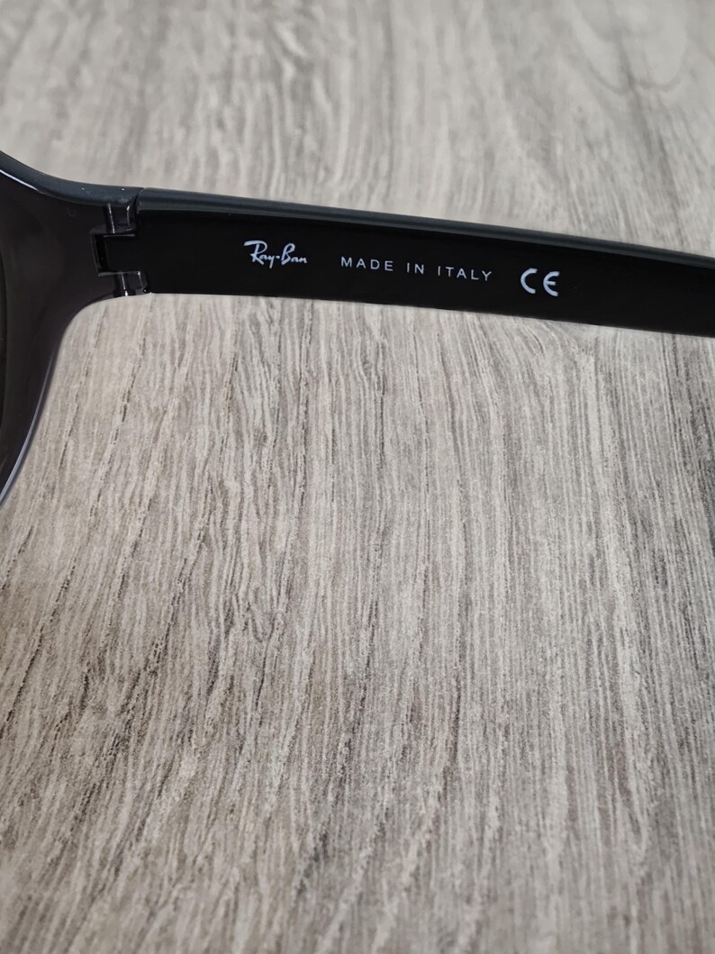 Puede incluir: Primer plano de la patilla de unas gafas de sol negras. El texto "Ray-Ban" est&aacute; impreso en blanco, junto con "MADE IN ITALY" y "CE". Las gafas de sol est&aacute;n sobre un fondo claro de vetas de madera.