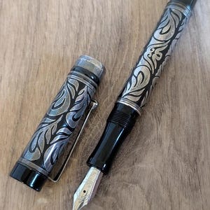 以下が含まれることがあります： 黒いボディとシルバーのアクセントが施された、詳細な万年筆。ペンは、バレルとキャップに装飾的で彫刻されたデザインが特徴です。ペン先はシルバーで、木製の表面に置かれています。
