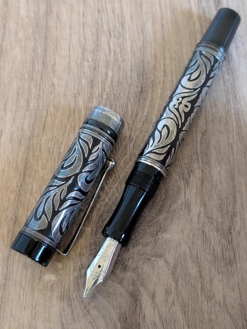 以下が含まれることがあります： 黒いボディとシルバーのアクセントが施された、詳細な万年筆。ペンは、バレルとキャップに装飾的で彫刻されたデザインが特徴です。ペン先はシルバーで、木製の表面に置かれています。