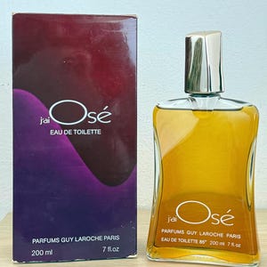 May include: A rectangular perfume box with a purple and dark red gradient, featuring the text J'ai Osé Eau de Toilette Parfums Guy Laroche Paris 200 ml 7 fl.oz. Beside it stands a clear glass bottle of amber liquid with a silver cap and matching text.