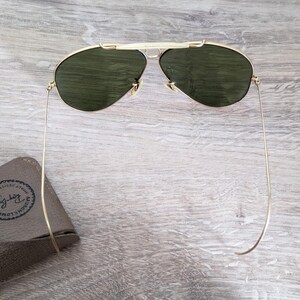 K&ouml;nnte beinhalten: Goldfarbene Sonnenbrille mit gr&uuml;nen Gl&auml;sern. Die Sonnenbrille hat ein klassisches Aviator-Design mit Doppelsteg und d&uuml;nnen B&uuml;geln. Ein braunes Etui mit Logo ist sichtbar.